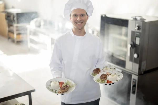 Glimlachende chef in uniform staand met kant-en-klare maaltijden in de keuken — Stockfoto