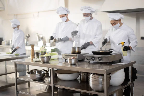 Team multirazziale di cuochi in uniforme e maschere per il viso che cucinano i pasti per un ristorante in cucina — Foto Stock