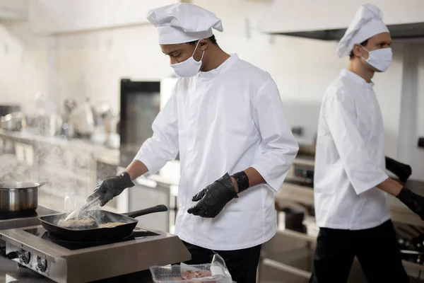 Due chef in uniforme e maschere per il viso che cucinano insieme in cucina professionale — Foto Stock