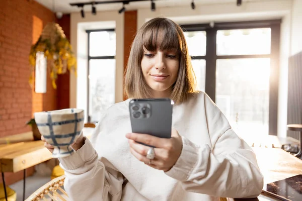 Wanita dengan telepon pintar dan cangkir kopi di ruang tamu cerah di pagi hari — Stok Foto