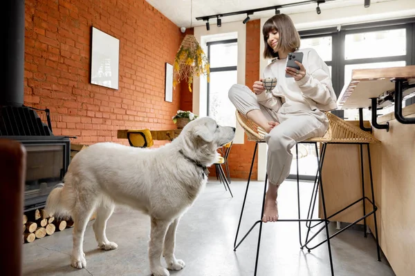 Donna seduta con una tazza di caffè e telefono al tavolo da bar con il suo cane in soggiorno soleggiato — Foto Stock