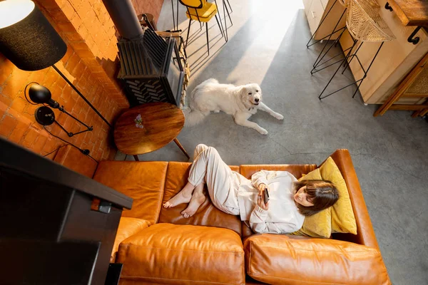 Kvinna ligger avslappnad på soffan och använder telefon med hund hemma — Stockfoto