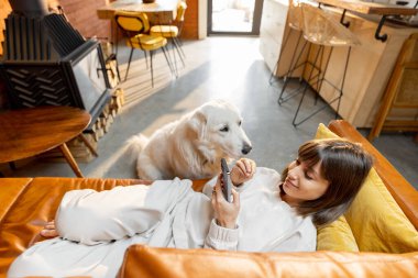 Kanepede rahatça uzanan, telefon kullanan, evde köpekle vakit geçiren bir kadın.