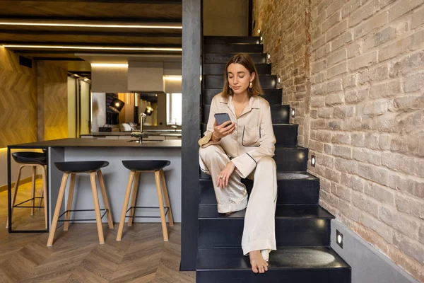 Mulher usando telefone enquanto sentado em escadas no apartamento moderno — Fotografia de Stock