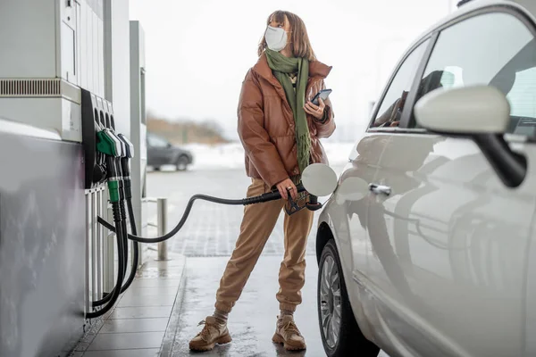 Женщина в маске для лица заправляет машину бензином с помощью смартфона — стоковое фото