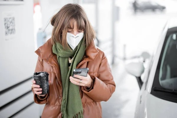 Жінка в масці для обличчя тримає смартфон і чашку кави під час заправки автомобіля на АЗС . — стокове фото