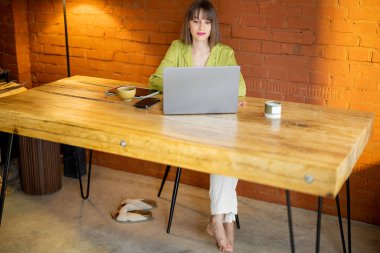 Kadın rahat evdeki ahşap masada otururken dizüstü bilgisayarla çalışıyor.