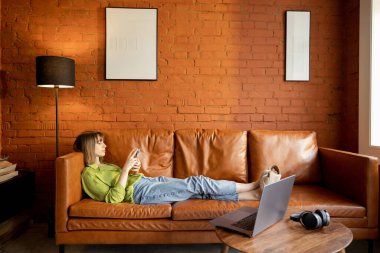 Evde deri bir koltukta dinlenirken telefonu kullanan bir kadın.