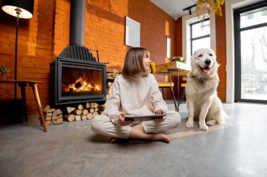Kadın köpeğiyle şöminenin yanında rahatça oturup dijital tablet kullanıyor.