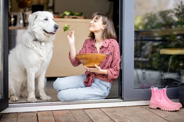 Lycklig kvinna äter sallad medan hon sitter med sin bedårande stora vita hund vid fönstret — Stockfoto