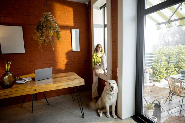Kvinnen som sitter i vinduskarmen og ser ut av vinduet med sin hvite, nydelige hund hjemme – stockfoto