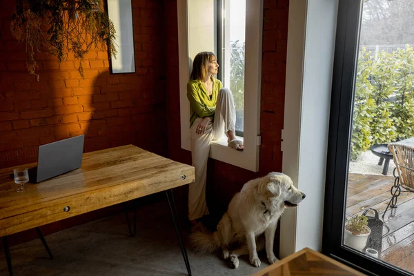 女人坐在窗台上，和她那只可爱的白狗呆在家里，望着窗外 — 图库照片