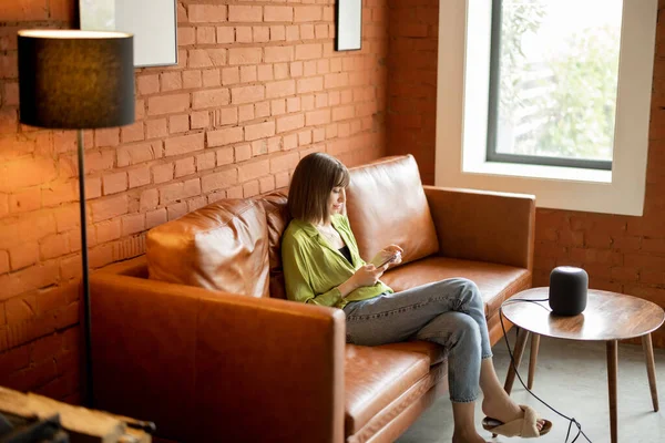 Žena s telefonem a inteligentní reproduktor sedí uvolněně doma — Stock fotografie