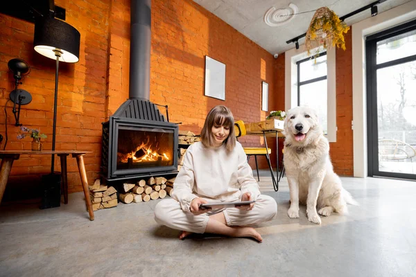 女人坐在壁炉边的地板上，和她的狗一起放松下来，在家里用数码平板电脑 — 图库照片