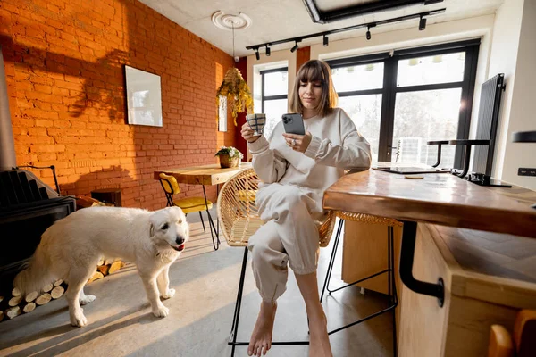 在阳光灿烂的客厅里，女人和她的狗一起坐在咖啡杯旁，打电话到吧台前 — 图库照片