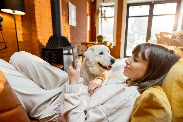 女人躺在沙发上，用电话休息，和狗呆在家里 — 图库照片