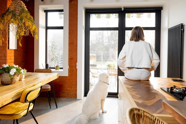 Kadın mutfak masasında tek başına oturuyor ve evdeki pencerelerden dışarı bakıyor.