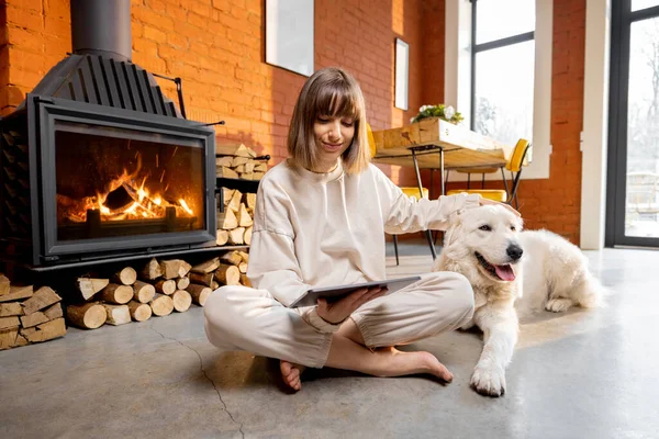 女人坐在壁炉边的地板上，和她的狗一起放松下来，在家里用数码平板电脑 — 图库照片