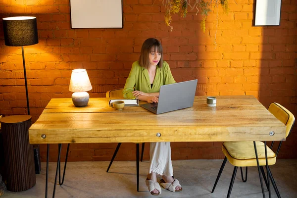 Kadın rahat evdeki ahşap masada otururken dizüstü bilgisayarla çalışıyor.