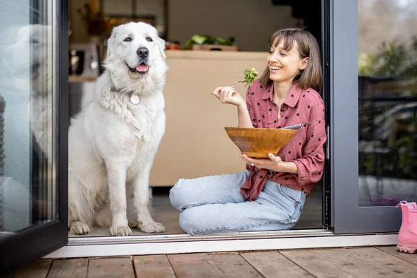 Mutlu kadın, sevimli beyaz köpeğiyle pencerede otururken salata yiyor.