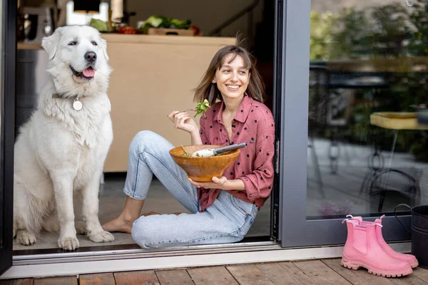 Ευτυχισμένη γυναίκα τρώει σαλάτα, ενώ κάθεται με το αξιολάτρευτο τεράστιο λευκό σκυλί της στο παράθυρο — Φωτογραφία Αρχείου