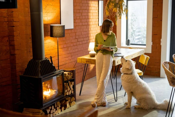 Kadın rahat oturma odasında köpeğiyle dikilirken dijital tabletle çalışıyor.