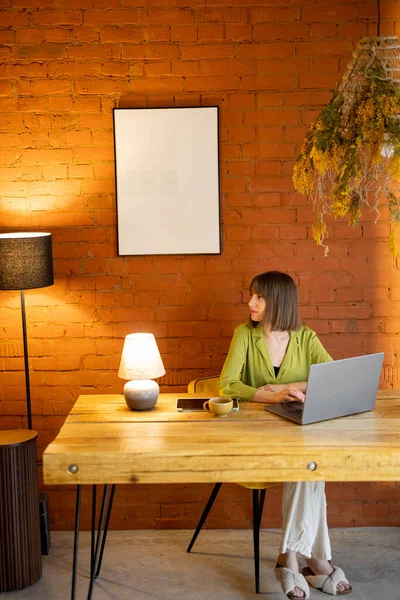 Kadın rahat evdeki ahşap masada otururken dizüstü bilgisayarla çalışıyor.