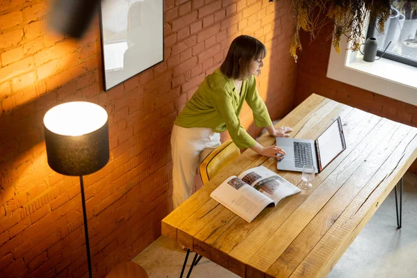 Kadın rahat oturma odasındaki ahşap masada otururken dizüstü bilgisayarla çalışıyor.