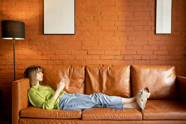 自宅の革のソファでリラックスしながら電話を使用して女性 — ストック写真