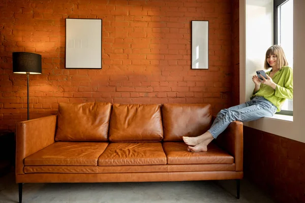 Kadın pencere eşiğinde oturuyor ve evde telefon kullanıyor.