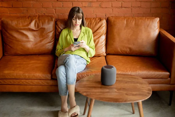 Donna con telefono e altoparlante intelligente seduta rilassata a casa — Foto Stock