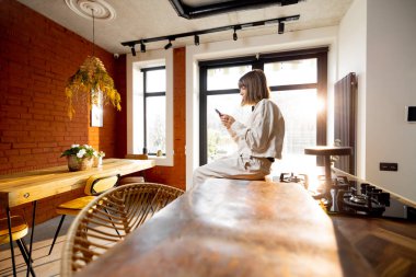 Evde tek başına mutfak masasında oturan bir kadın telefonu kullanıyor.