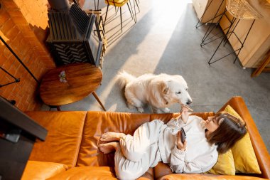 Kanepede uzanan ve evde köpekle telefon kullanan bir kadın.