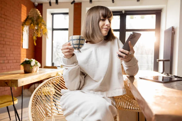 Mujer con teléfono inteligente y taza de café en la sala de estar soleada por la mañana — Foto de Stock