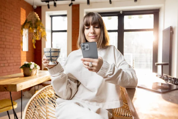 早上在阳光灿烂的客厅里拿着智能手机和咖啡杯的女人 — 图库照片