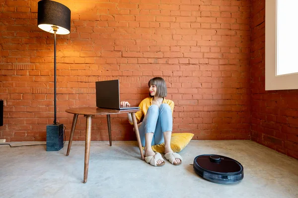 Mulher trabalhar no laptop enquanto aspirador robótico limpeza chão em casa — Fotografia de Stock