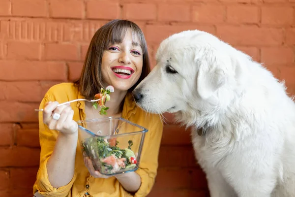 Retrato de una joven mujer alegre sentada con su lindo perro blanco y comiendo ensalada saludable —  Fotos de Stock