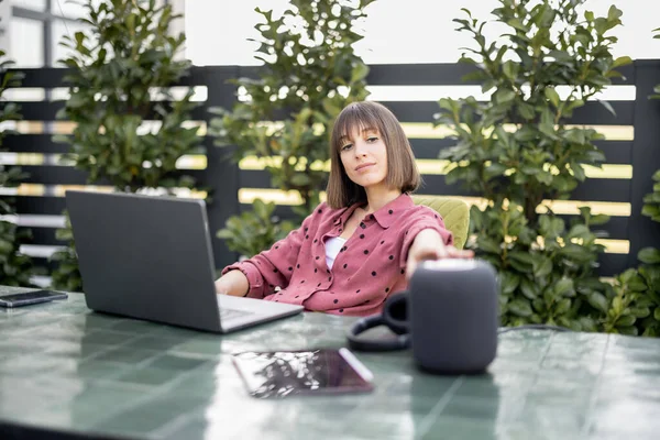 Woman touching smart speaker while sitting by the table and works in the garden — Stock Photo, Image