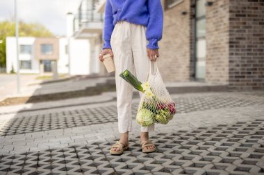 Woman with mesh bag full of vegetables and reusable coffee cup outdoors