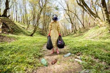 Gaz maskeli ve koruyucu elbiseli bir kadın ormanda dağınık plastik çöpleri topluyor.