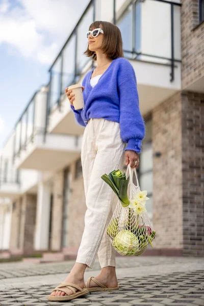 Woman walks with mesh bag full of vegetables and reusable coffee cup near home outdoors — Φωτογραφία Αρχείου