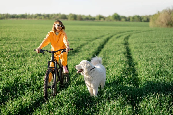 Mulher monta uma bicicleta em um campo com cão — Fotografia de Stock