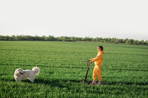 Woman rides electric scooter on green field with a dog — Photo
