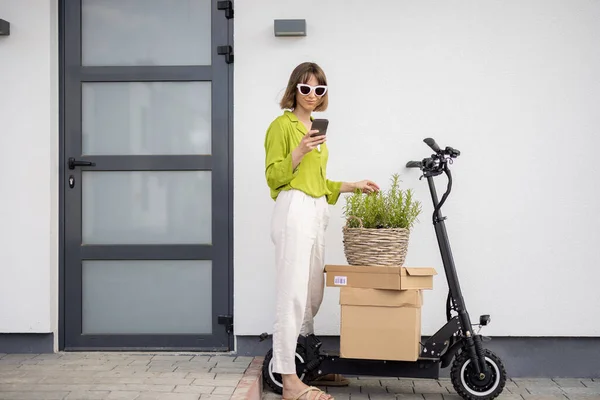 Telefonu, elektrikli scooter 'ı ve eve giden paketleri olan bir kadın. — Stok fotoğraf