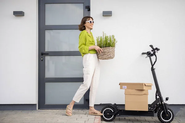 Woman carrying basket with herbs on a porch of her house with electric scooter and parcels nearby — Stockfoto