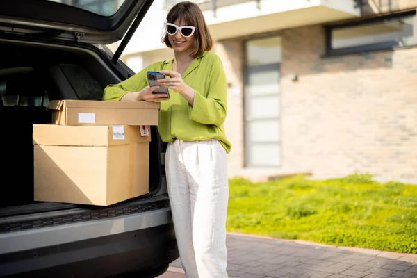 Kadının biri elinde paketlerle arabanın bagajında dikilirken telefonu kullanıyor.