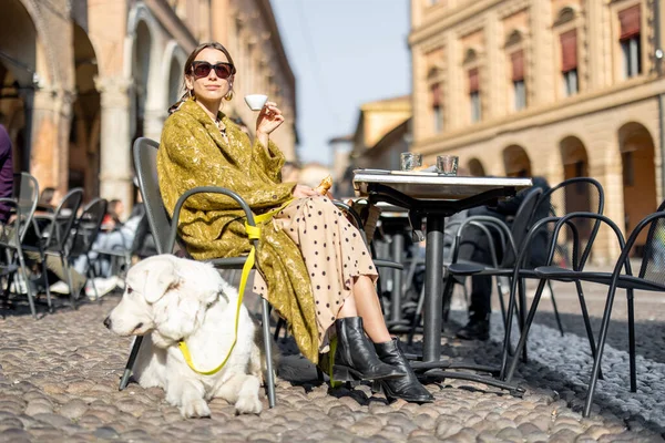 Beyaz köpeğiyle birlikte eski Bologna şehrindeki açık kafede oturan bir kadın.