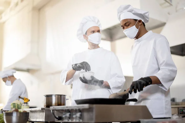 Equipo multirracial de cocineros en uniforme y mascarillas para cocinar comidas para un restaurante en la cocina —  Fotos de Stock