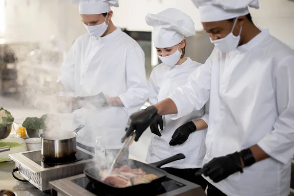 Многорасовая команда поваров в униформе и масках для лица готовит еду для ресторана на кухне — стоковое фото
