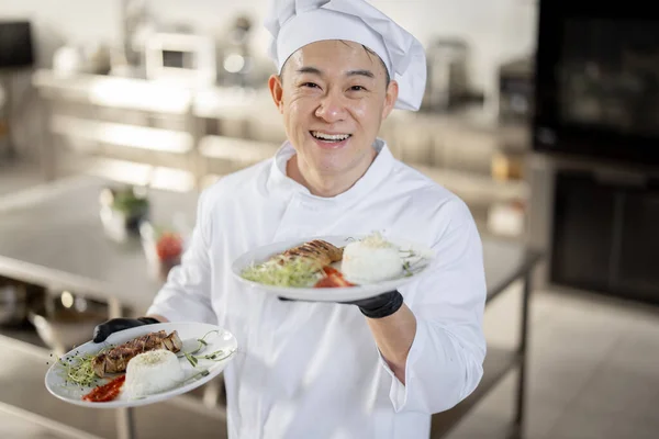 Portret van lachende chef-kok in uniform staand met kant-en-klaar maaltijd in de keuken — Stockfoto